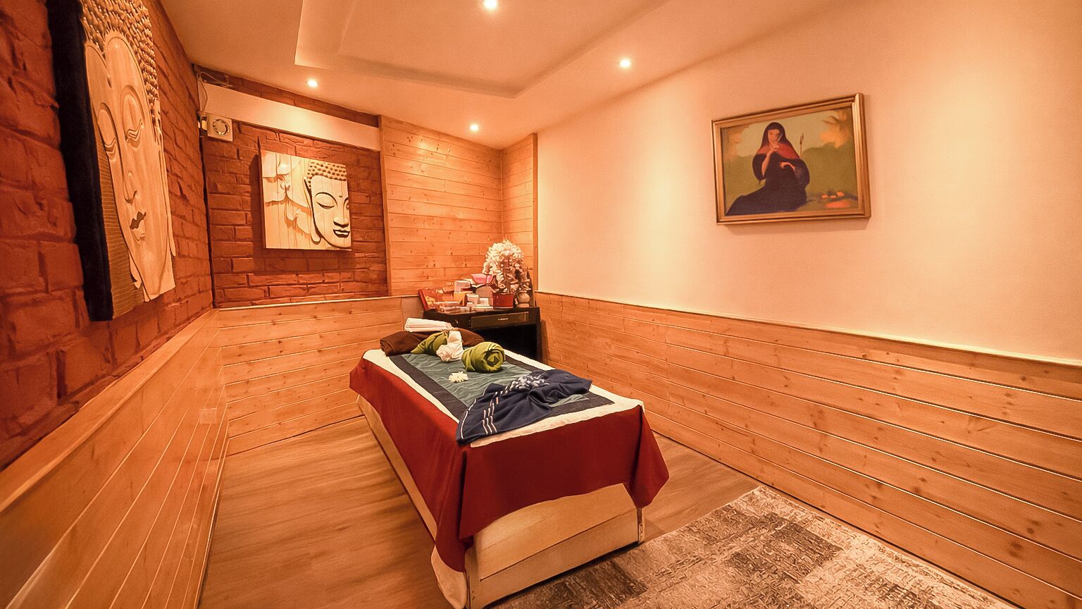 A room with wooden walls, a bed with a red blanket, a small table with a vase, and a painting on the wall.