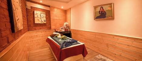 A room with wooden walls, a bed with a red blanket, a small table with a vase, and a painting on the wall.