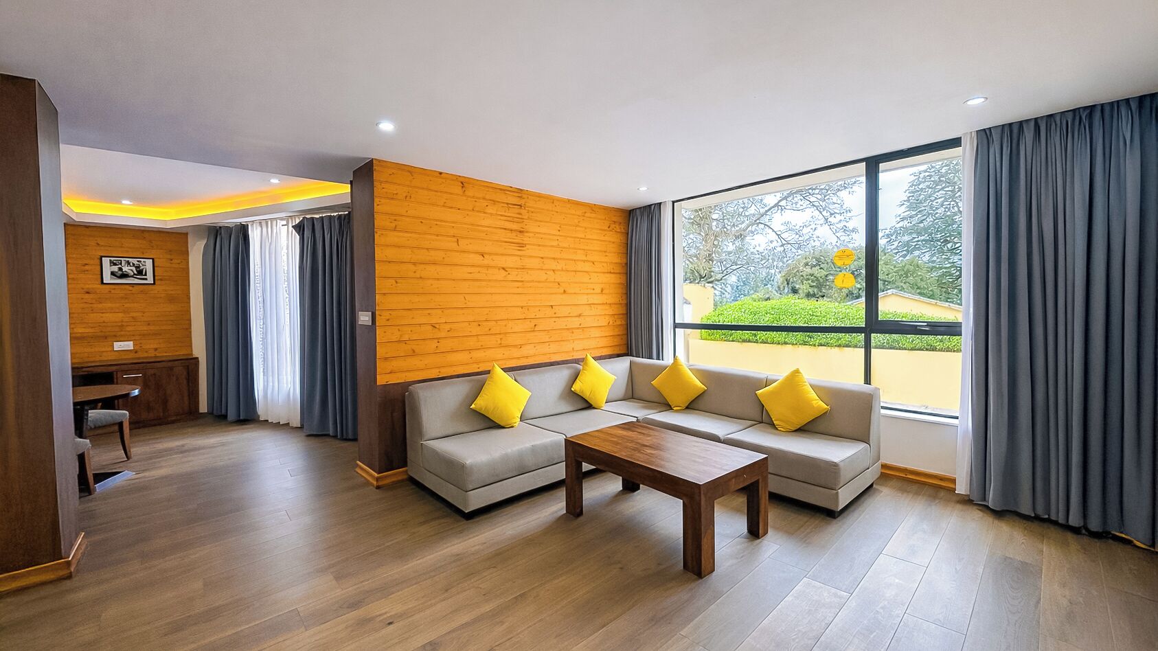 A modern living room with a wooden accent wall, a grey sofa set with yellow cushions, a wooden coffee table, and a large window with curtains.