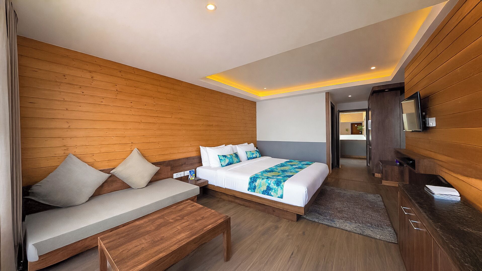 A modern hotel room with a wooden wall, a large bed with a blue and white blanket, a wooden bench, and a TV mounted on the wall.