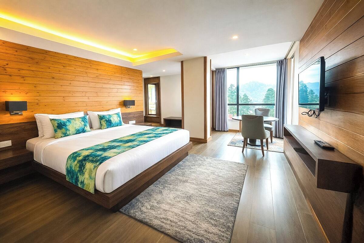 Una habitación de hotel moderna con cabecera de madera, una cama grande con manta azul y verde, un escritorio con silla y un televisor montado en la pared.