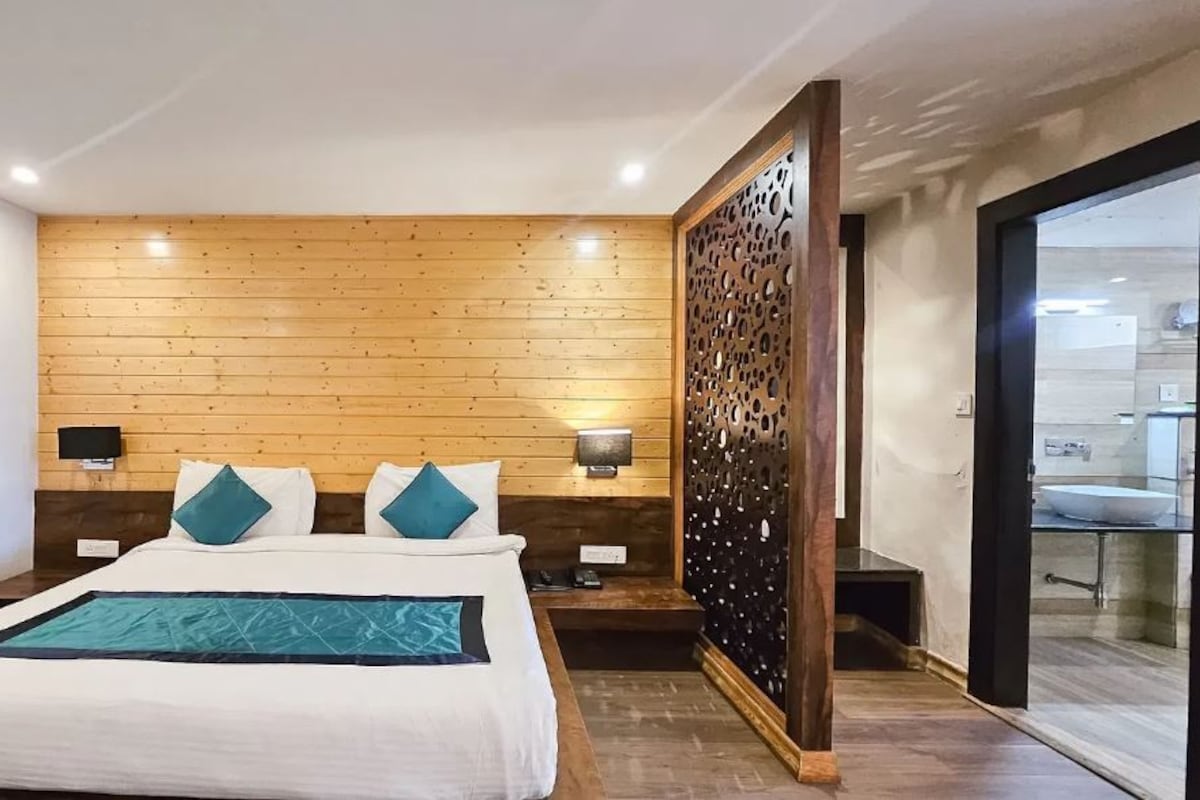 Un dormitorio moderno con cabecera de madera, una cama grande con ropa de cama blanca y detalles en color turquesa, una mesita de noche con una lámpara y vistas a una cocina a través de una puerta abierta.