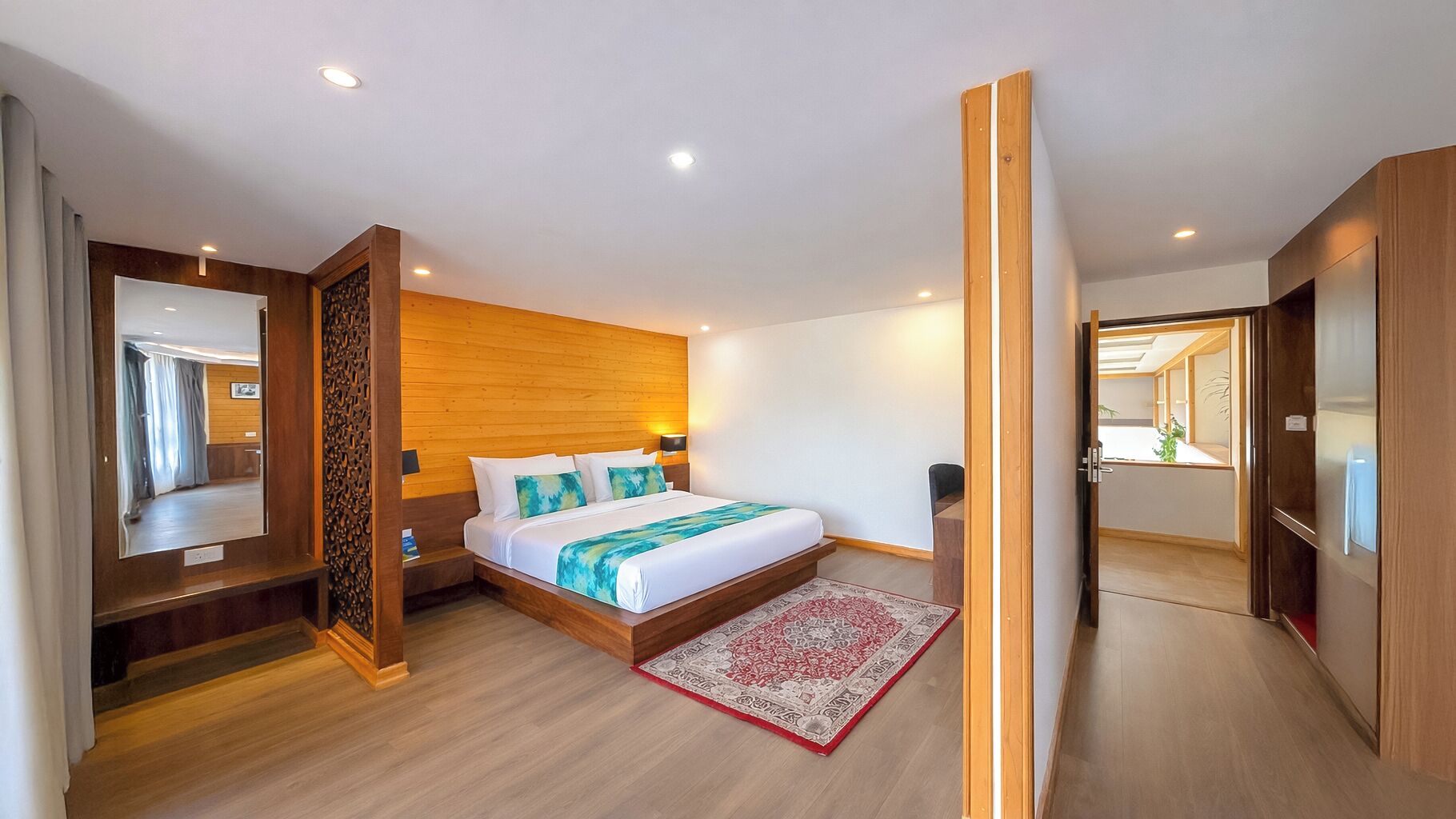 A modern hotel room with a wooden headboard, a large bed with white linens, a red and beige patterned rug, and a wooden floor.