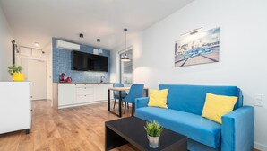 A modern living room with a blue sofa, a coffee table with a plant, and a kitchen area with blue tiles and a flat-screen TV. - MARGI Figo Apartament (Szczecin)