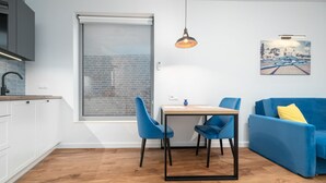 A modern kitchen with white cabinets, a wooden countertop, and a window with a view of a brick wall. There is a dining table with blue chairs and a blue sofa with a yellow pillow. - MARGI Figo Apartament (Szczecin)