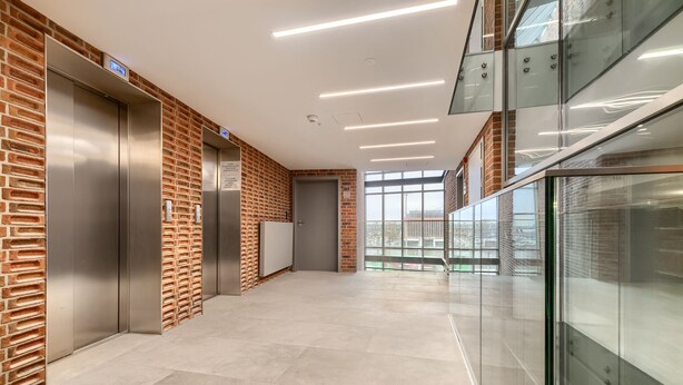A modern elevator area with brick walls and glass railings, featuring a clean, tiled floor and overhead lighting. - MARGI Figo Apartament (Szczecin)
