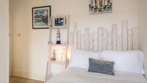 A bedroom with a white bed, a headboard made of wooden planks, a small shelf with decorative items, and a painting of sailboats on the wall. - Beach in Reach - group house for up to 14 with sea views (Eastbourne)