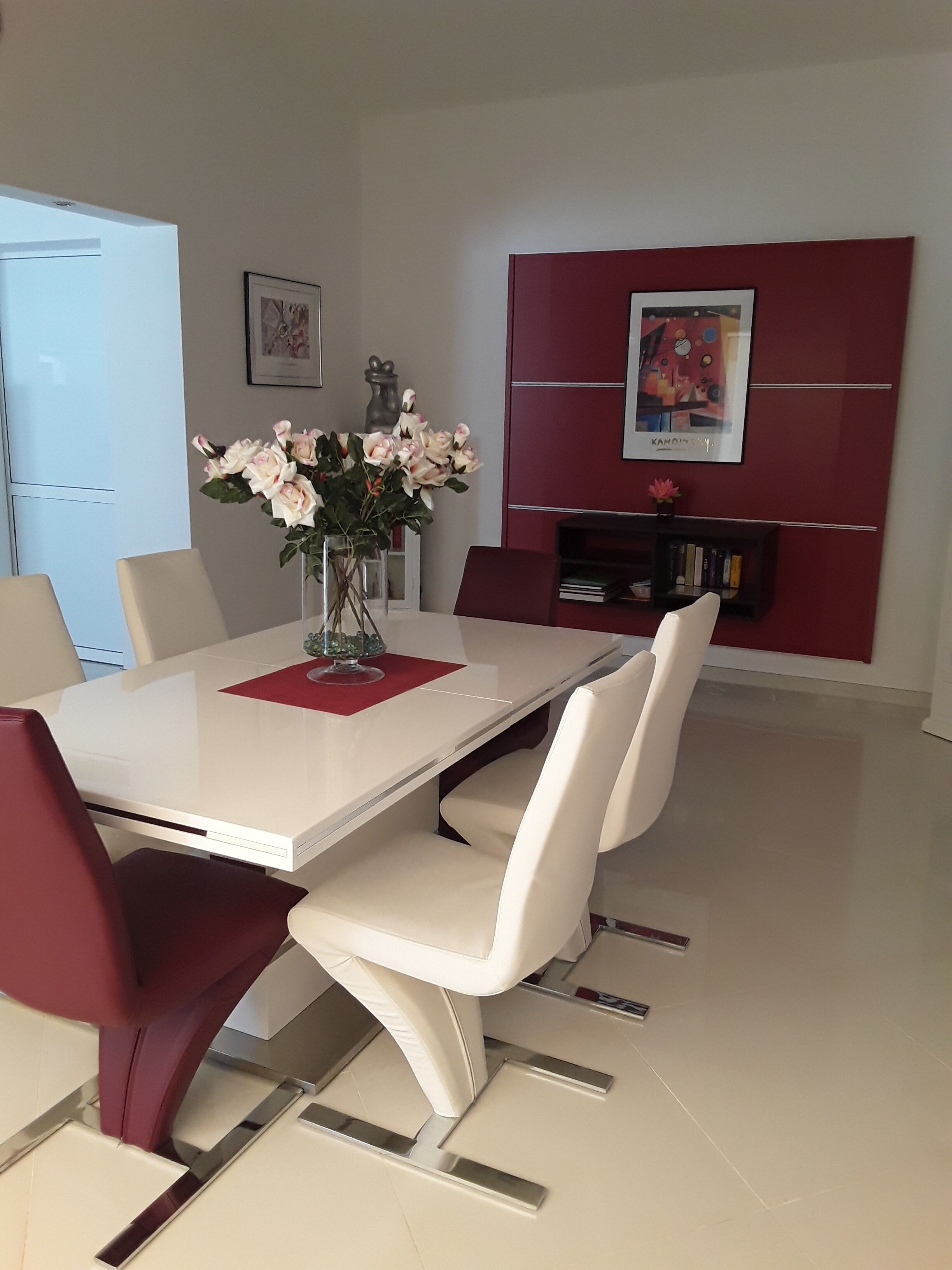 A dining area with a white table, red and white chairs, a vase with flowers, and a bookshelf with books and a framed picture.
