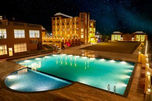A swimming pool with a night sky view, illuminated buildings, and a well-lit deck area. - The Nangal Resort (Udaipurwati)