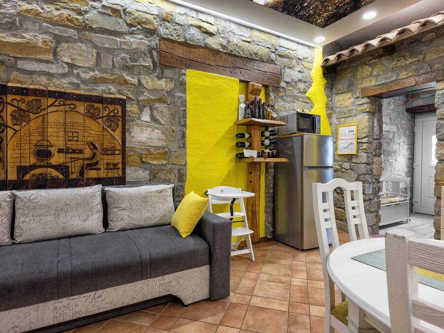 A cozy living area with a stone wall, a sofa with patterned cushions, a small white table, and a kitchen area with a refrigerator and wine bottles.