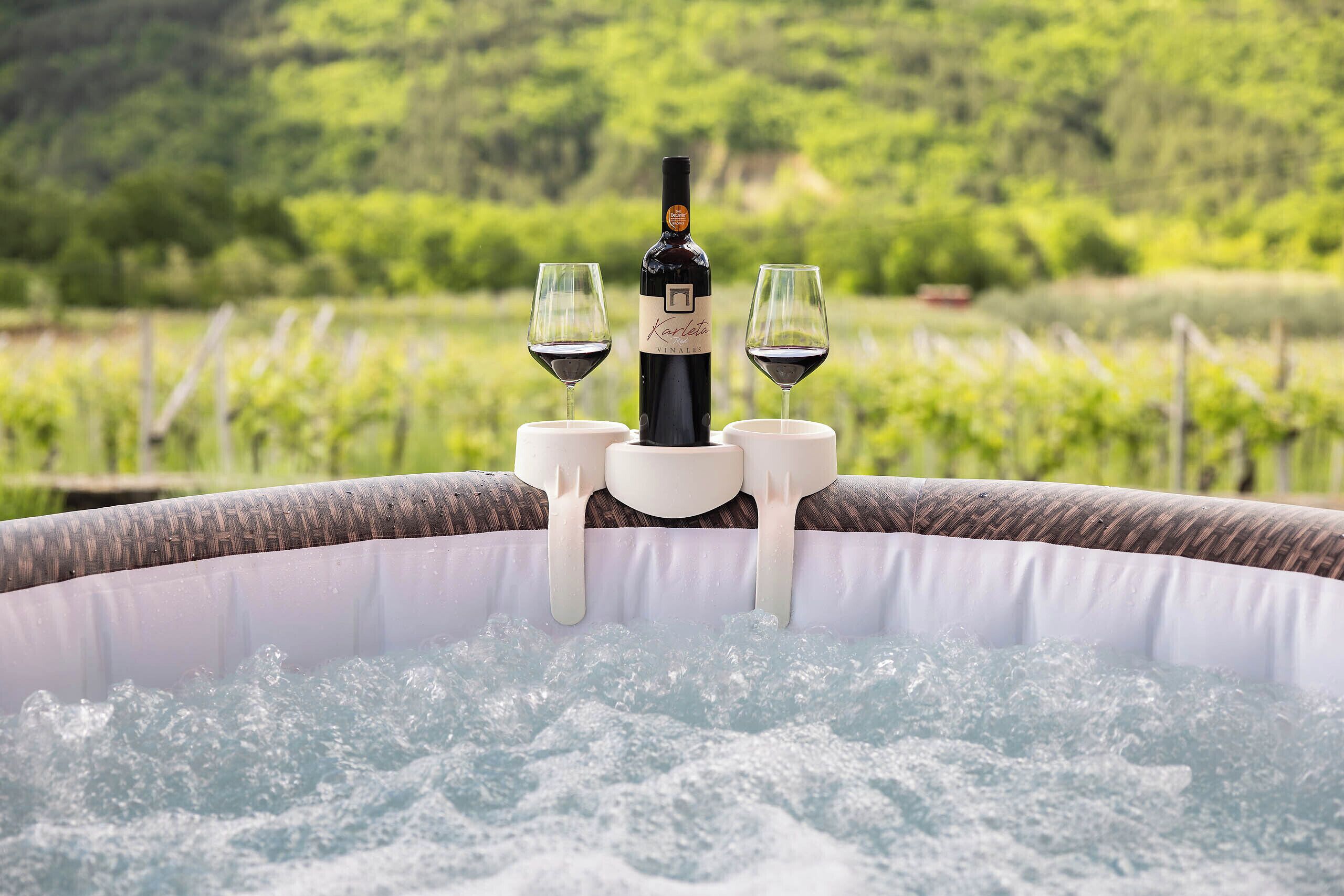 A hot tub with a bottle of wine and two glasses, surrounded by a lush green vineyard.