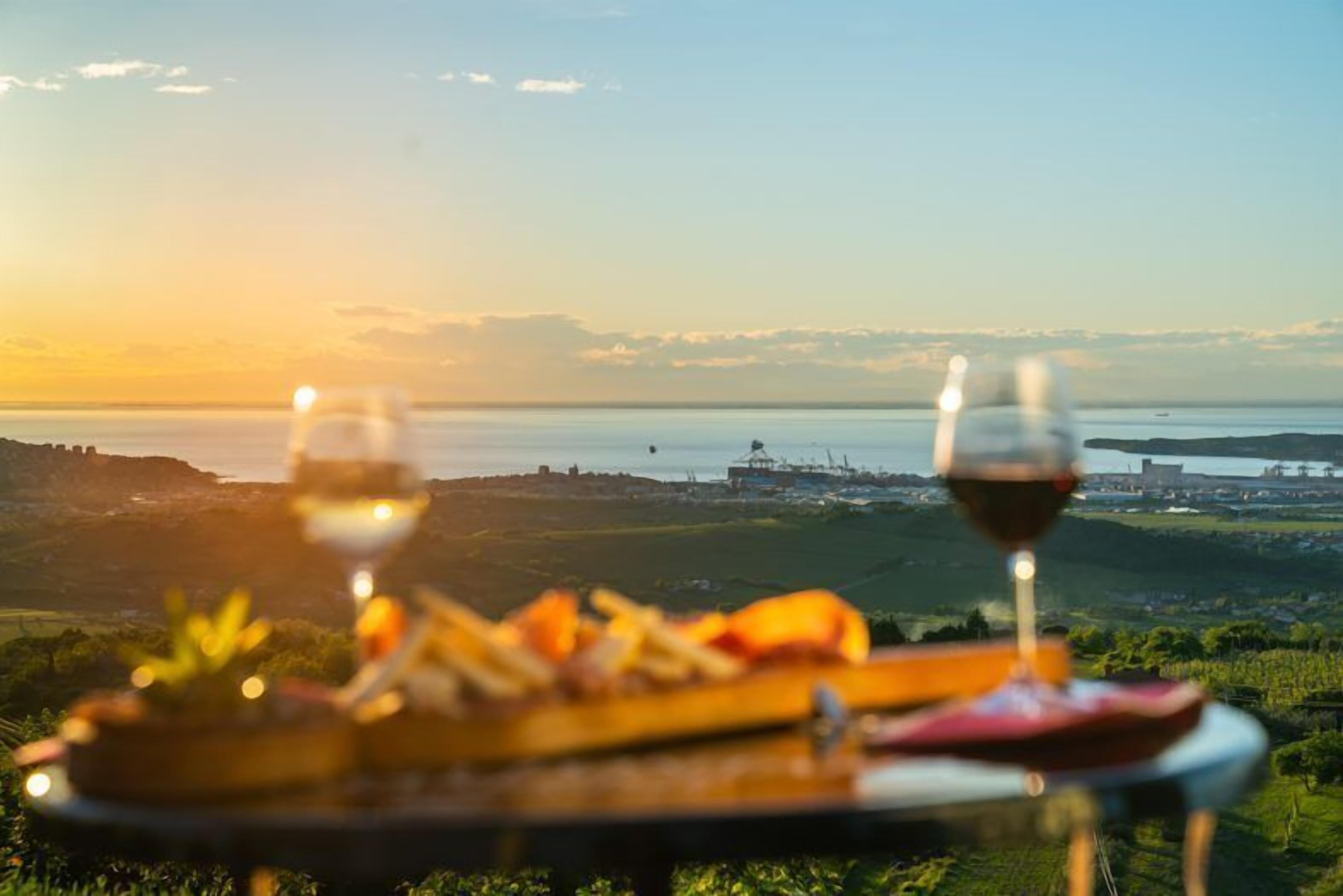 A table with a glass of red wine and a plate of food, overlooking a scenic landscape with hills and a body of water.