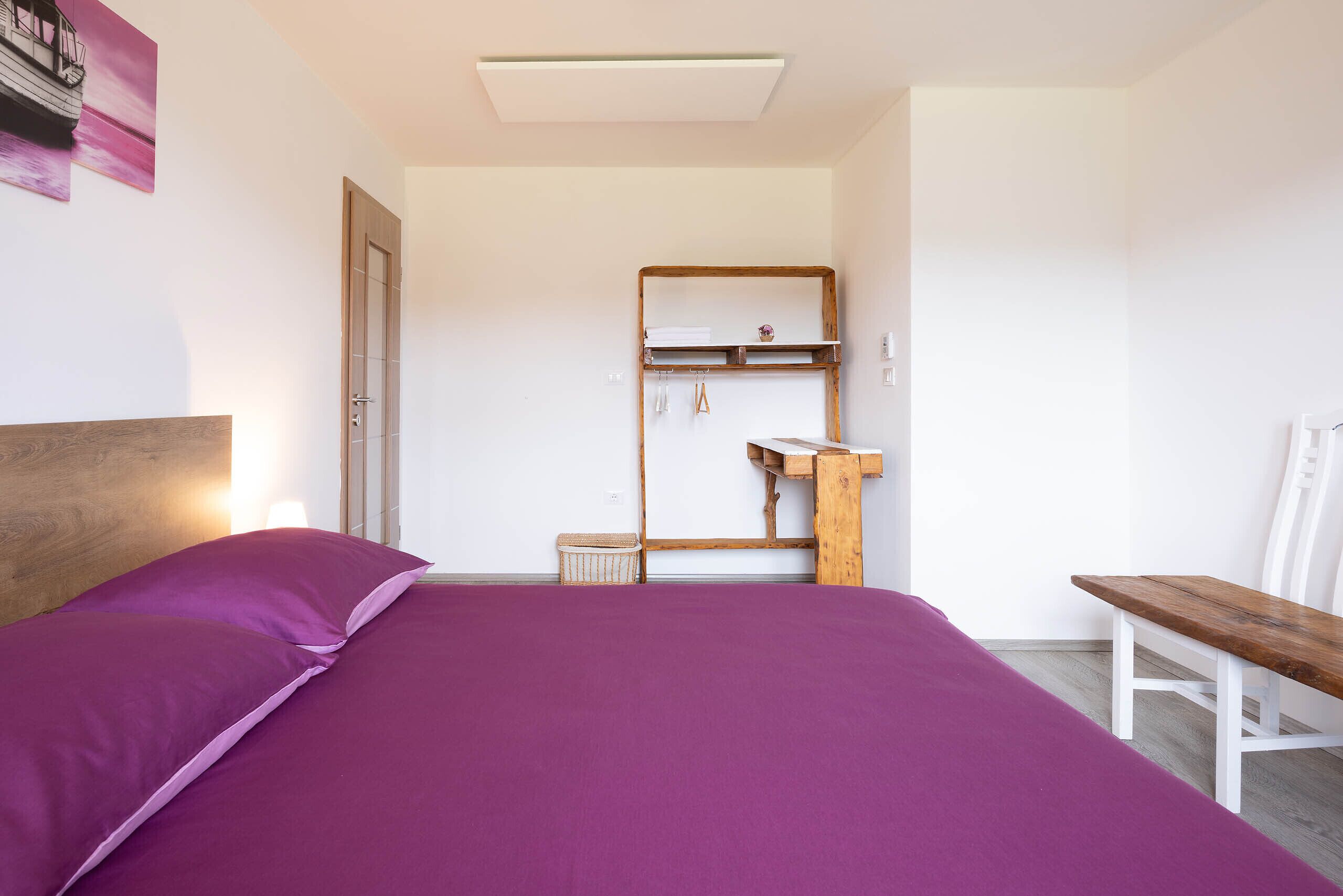 A bedroom with a bed, a wooden headboard, a small wooden bench, a white chair, a wooden shelf with hanging items, and a door.