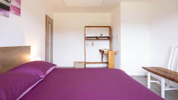 A bedroom with a bed, a wooden headboard, a small wooden bench, a white chair, a wooden shelf with hanging items, and a door.