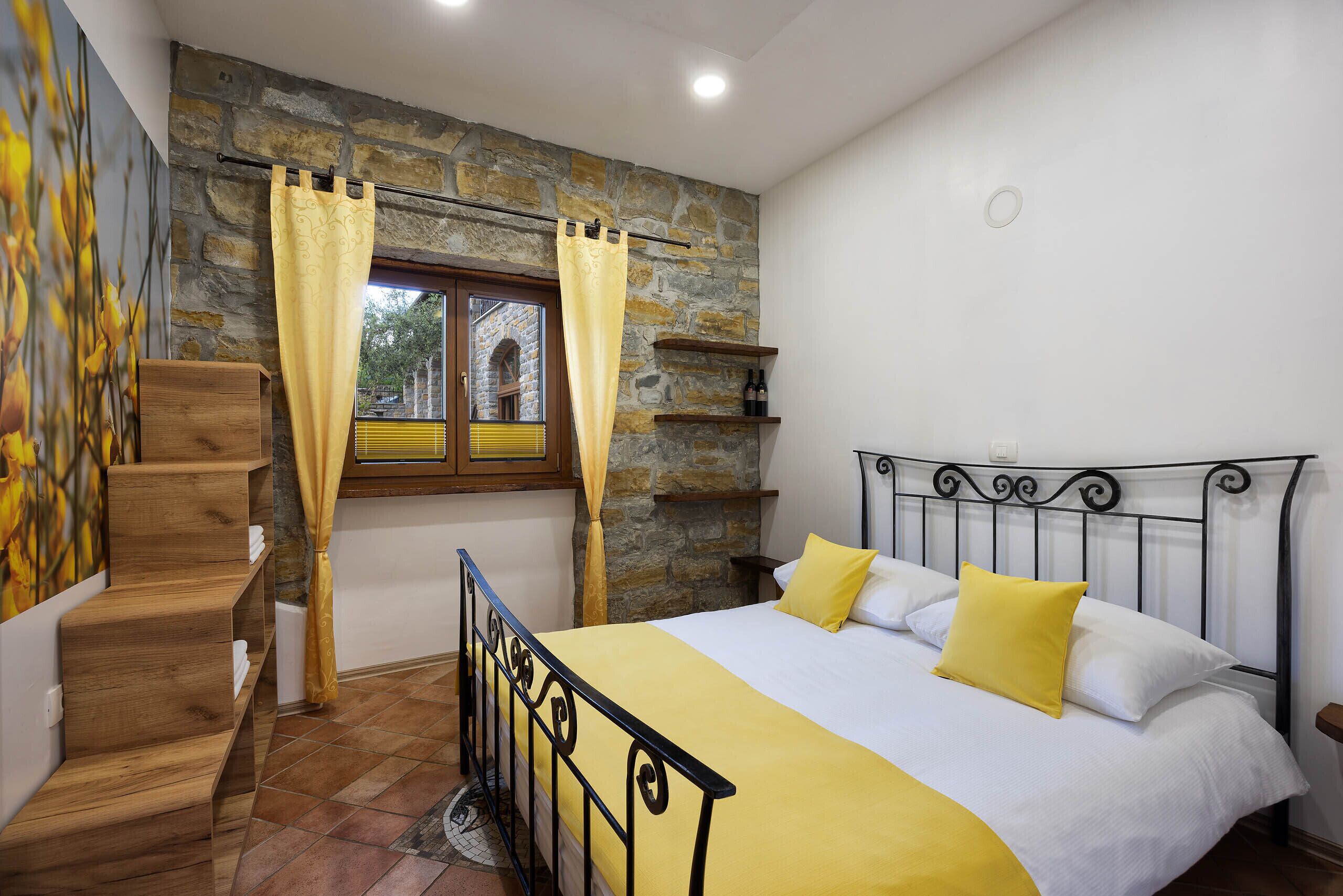 A bedroom with a stone wall, a bed with yellow bedding, a wooden staircase, and a window with curtains.