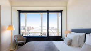 Une chambre d’hôtel avec un grand lit, deux lampes de chevet, une chaise et une vue sur les bâtiments par une grande fenêtre.