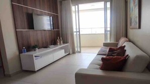 A modern living room with a white sofa, a flat-screen TV on a wooden TV stand, and a sliding glass door leading to a balcony. - Apto 2/ Q próximo ao Hospital premium (Goiania)
