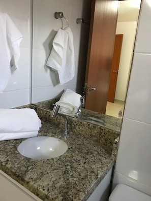 Um banheiro com bancada de granito, pia branca e porta-toalhas com várias toalhas brancas.