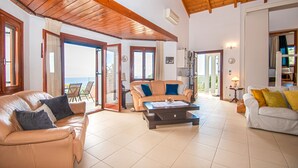 A spacious living area with a wooden ceiling, a beige sofa, a white sofa, and a coffee table. There is a sliding glass door leading to an outdoor area. - DN Beach Coast Villa (Zakynthos)