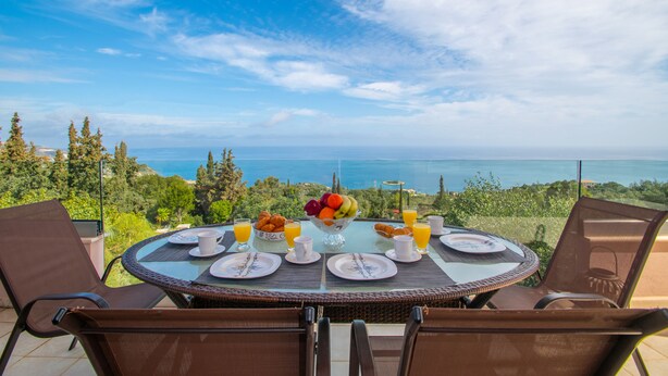 A round dining table with a glass top, surrounded by brown chairs, set for breakfast with plates, cups, and a fruit bowl. - DN Beach Coast Villa (Zakynthos)