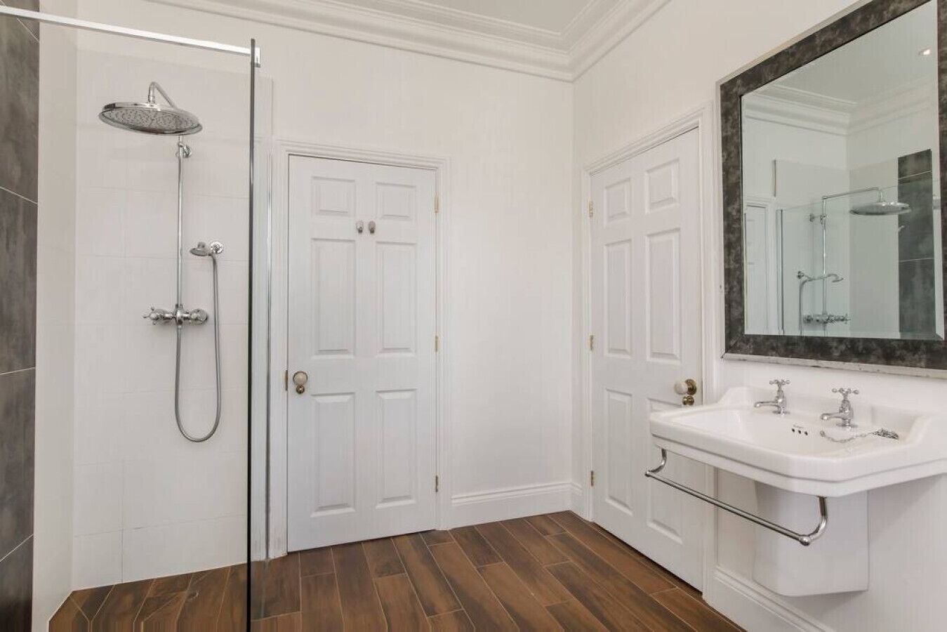 A bathroom with a shower, two white doors, a sink with a mirror, and wooden flooring.