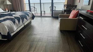 Ein Hotelzimmer mit einem großen Bett, einem Sessel, einem Schminktisch und Meerblick durch die Balkontüren.
