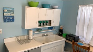 A kitchen with white cabinets, a sink, a countertop, and a window with a white curtain. - Beach Side Studio #1 Apts by RedAwning (Norfolk)