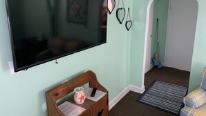 A living room with a flat-screen TV mounted on the wall, a wooden bench with a candle and remote control, a patterned rug, and a plaid armchair. - Beach Side Studio #1 Apts by RedAwning (Norfolk)