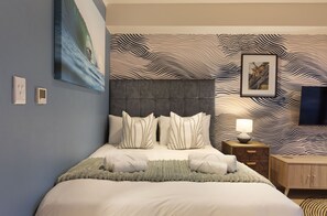 A bedroom with a large bed, a headboard, a bedside table with a lamp, and a wall with a wavy pattern. - The Tokyo 1508 (Cape Town)