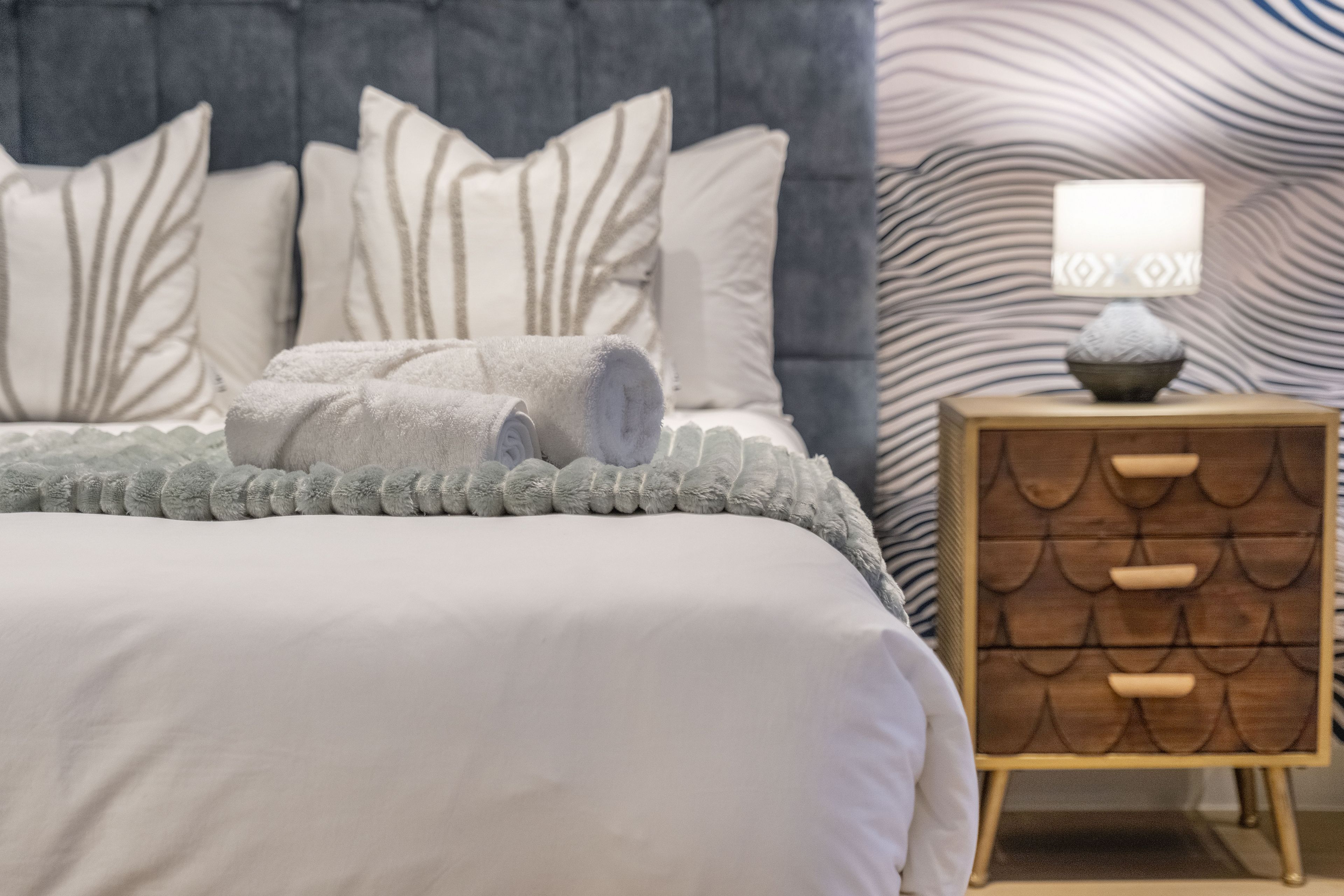 A neatly made bed with a grey blanket and rolled towels, a bedside lamp, and a wooden nightstand with drawers.