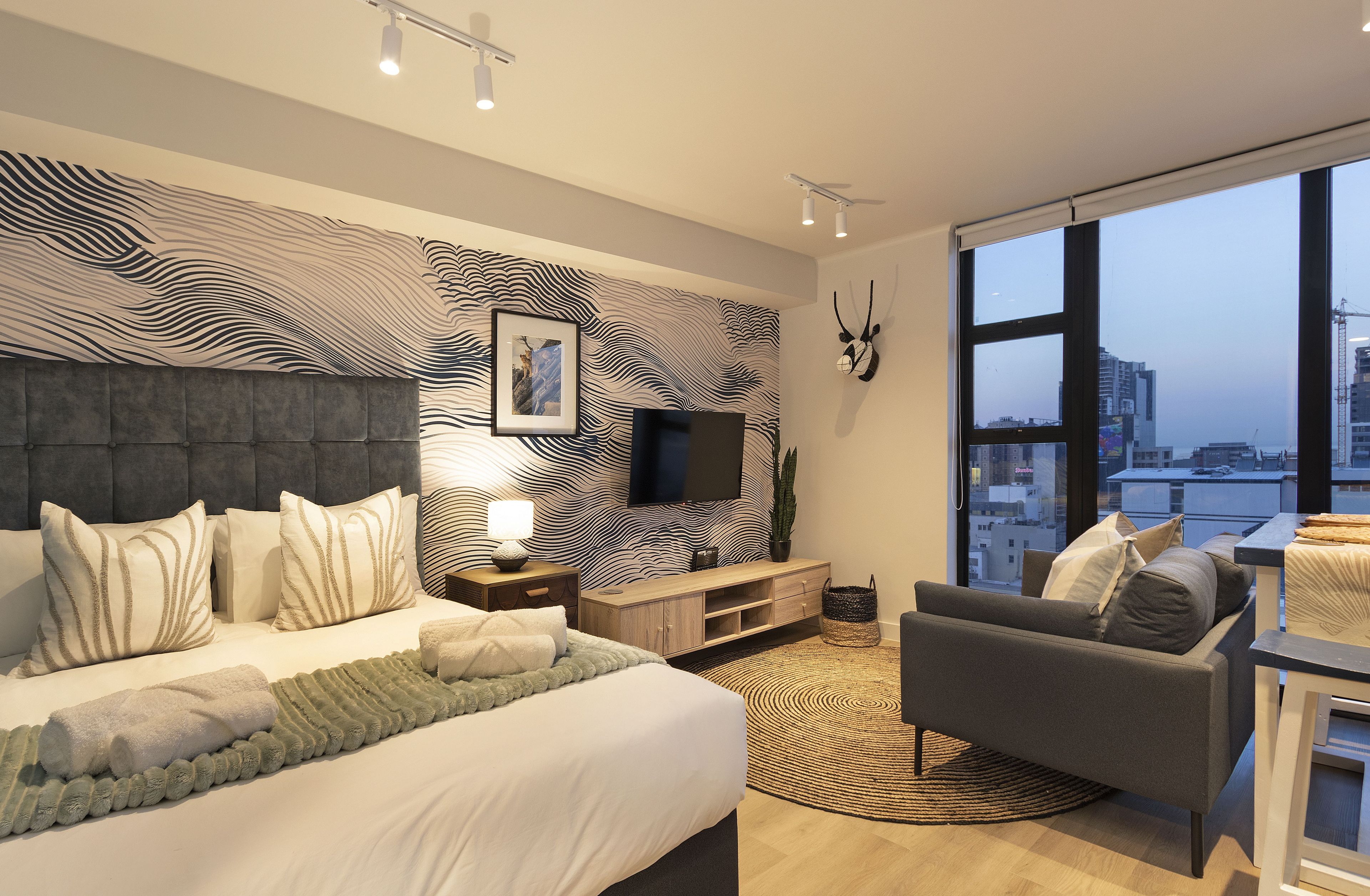 A modern bedroom with a large bed, a TV, a city view, and a cozy seating area.