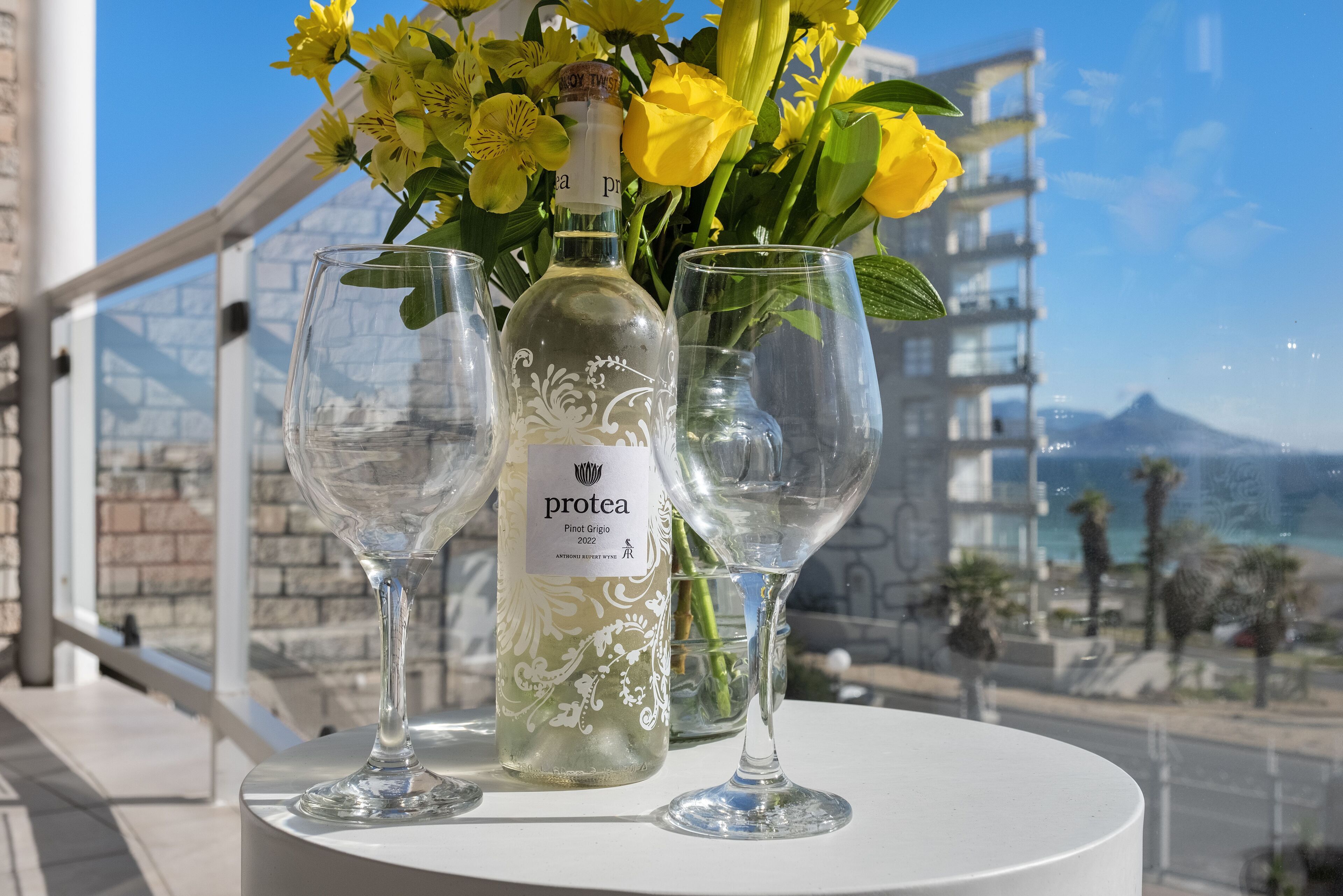 Una botella de Protea Pinot Grigio 2022, dos copas de vino vacías y un ramo de flores amarillas en un balcón con vistas a un edificio y al mar.