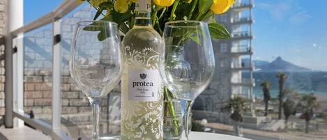 Una botella de Protea Pinot Grigio 2022, dos copas de vino vacías y un ramo de flores amarillas en un balcón con vistas a un edificio y al mar.