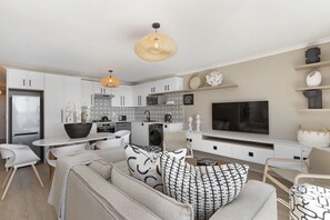 A modern kitchen with white cabinets, a dining area with a white table and chairs, a sofa with patterned cushions, and a large flat-screen TV mounted on the wall.