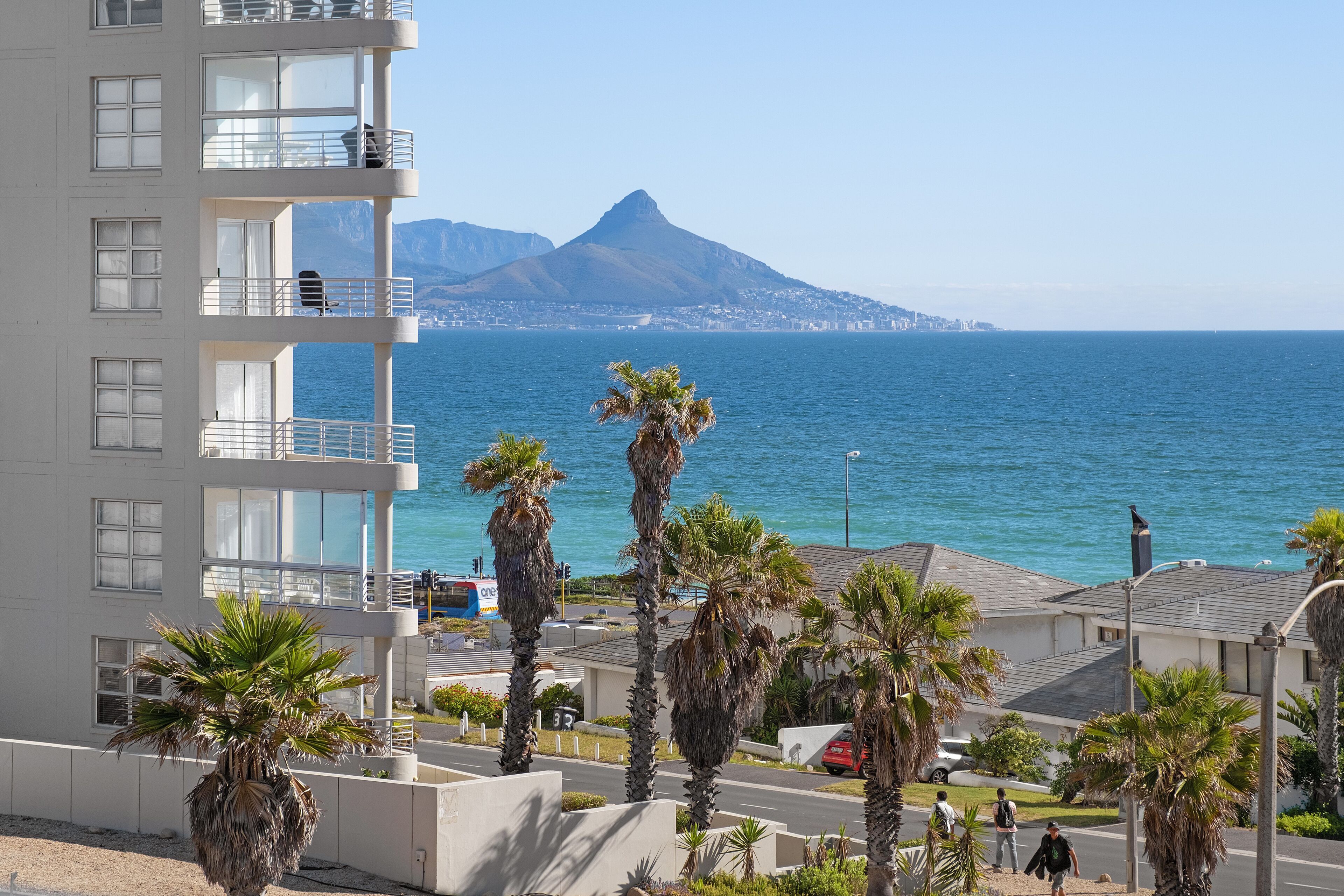A coastal building with balconies overlooking the sea, palm trees, and a distant mountain.