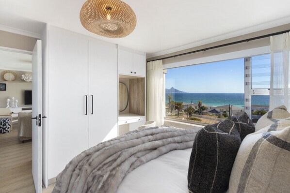 A bedroom with a large window, a bed with a fluffy blanket, and a view of the ocean.