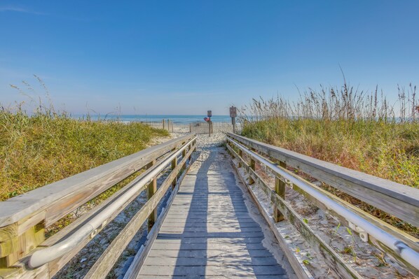 A wooden boardwalk leading to the beach, surrounded by tall grasses and a clear blue sky. - Beachfront Condo w/ Balcony & Pool in Myrtle Beach (Myrtle Beach)