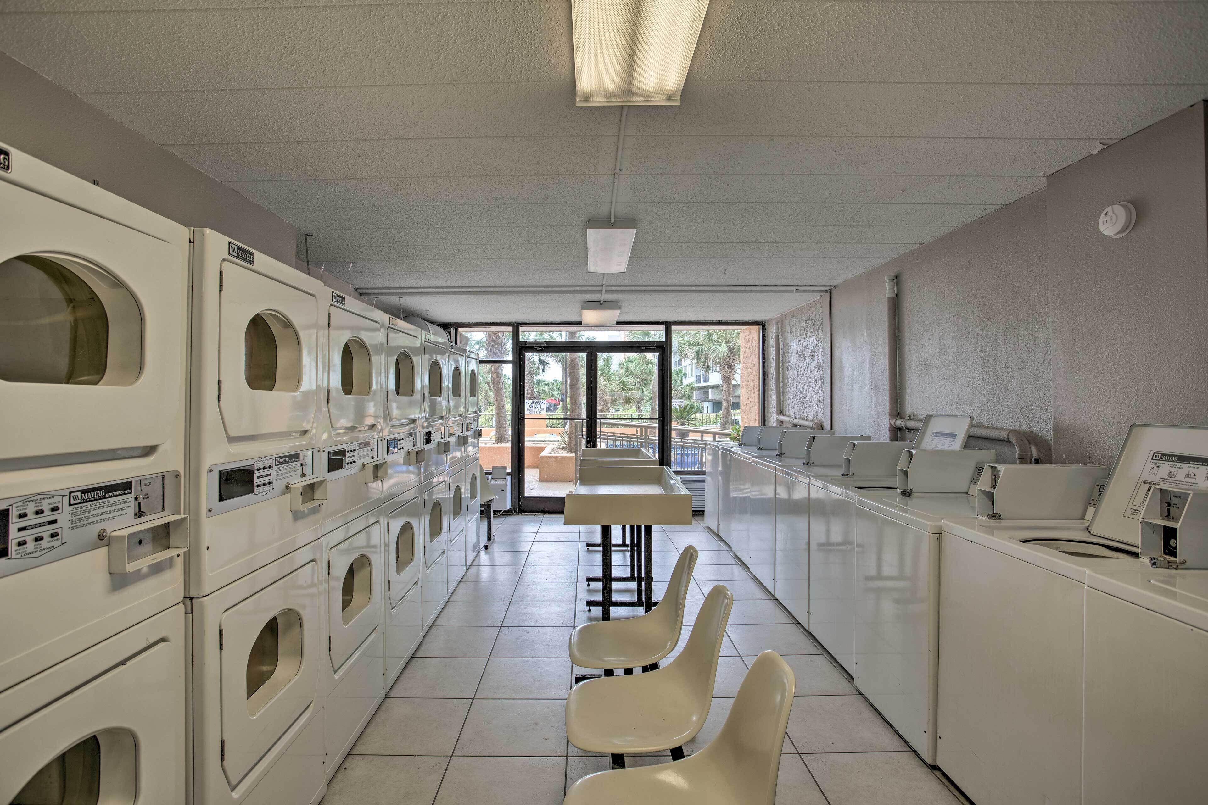A laundry facility with multiple washing machines and dryers, a long counter, and a view of the outdoors through large windows.
