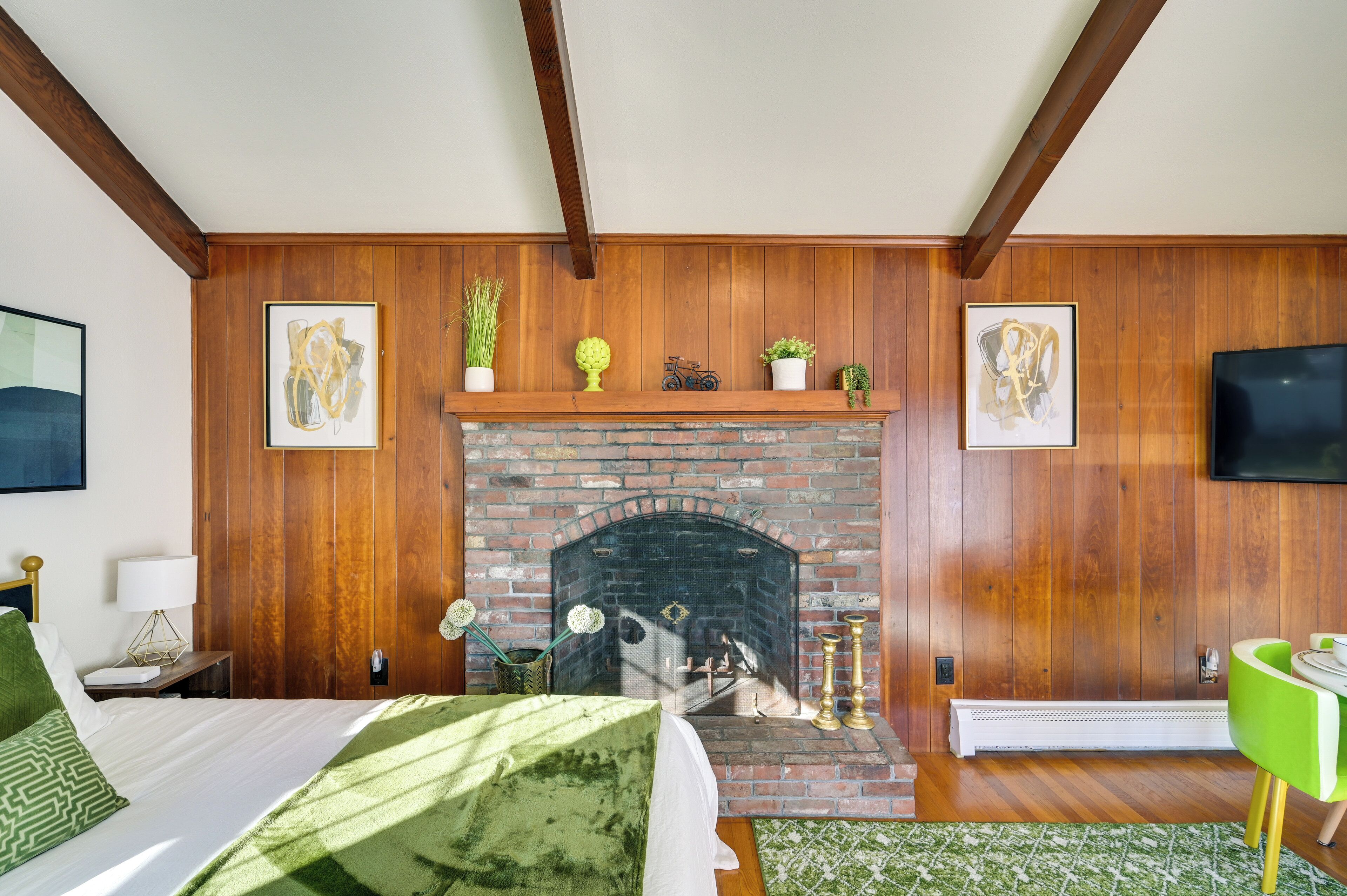 A room with a brick fireplace, wooden panel walls, a bed with green bedding, a green chair, and a flat-screen TV mounted on the wall.