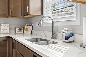 Una cocina moderna con dos lavamanos de acero inoxidable, armarios de madera, un ventanal con persianas y decoración inspiradora en la pared.