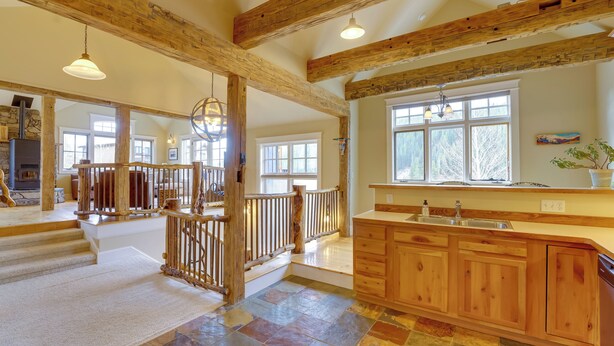 A spacious kitchen with wooden cabinets and a stone tile floor, a wooden staircase, and large windows allowing natural light.