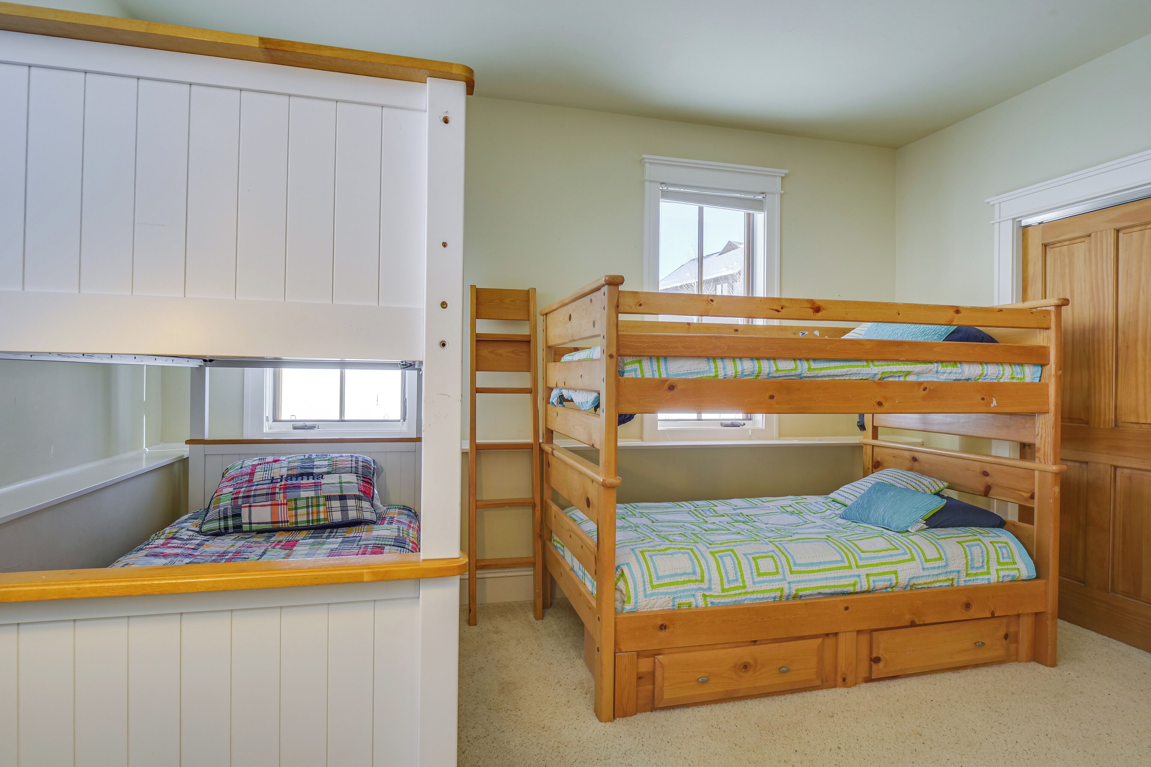 A bunk bed with a desk and ladder, a window, and a door in a room with light-colored walls.