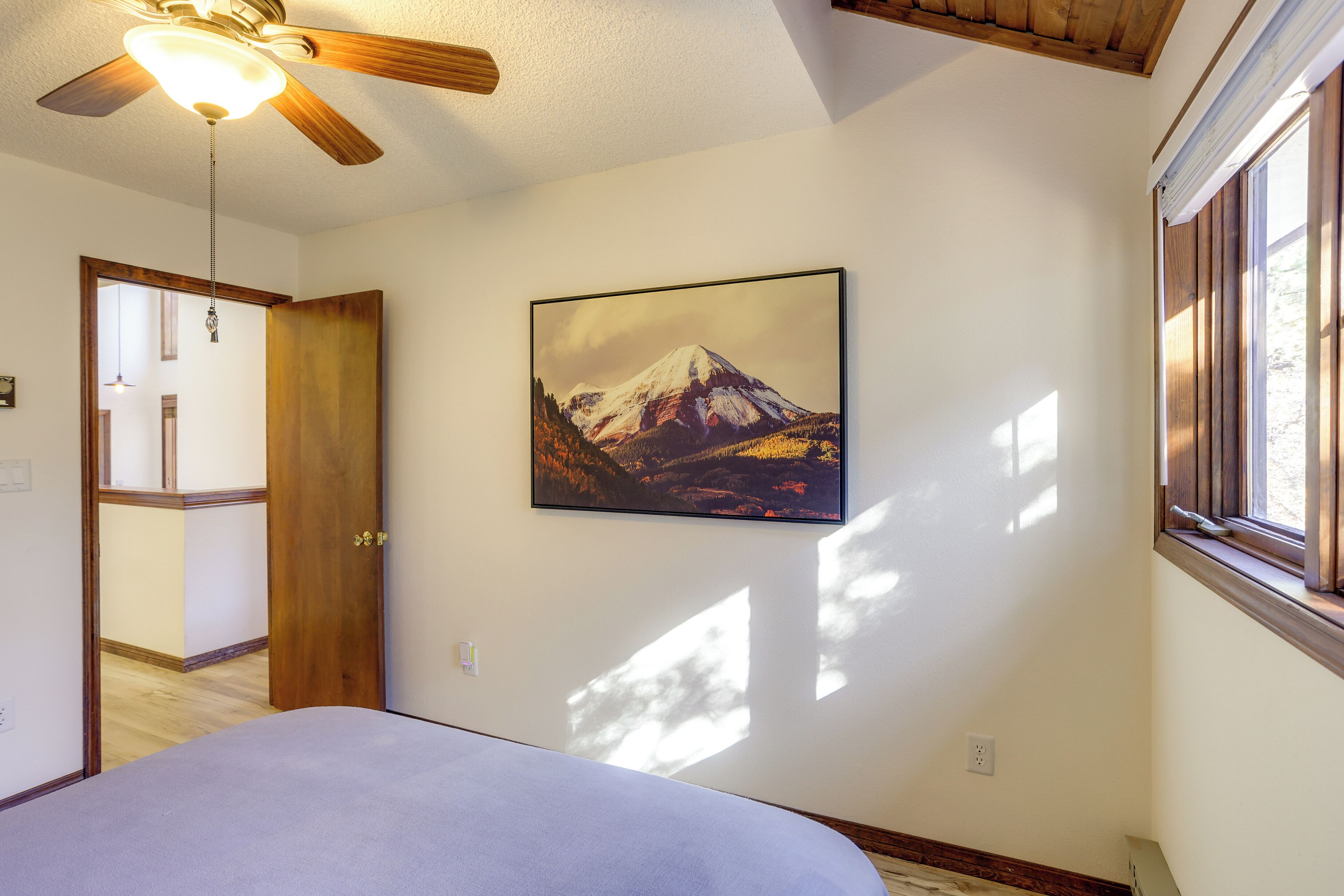Una camera da letto con un letto, una ventola a soffitto, un quadro incorniciato di una montagna e una finestra con persiane.