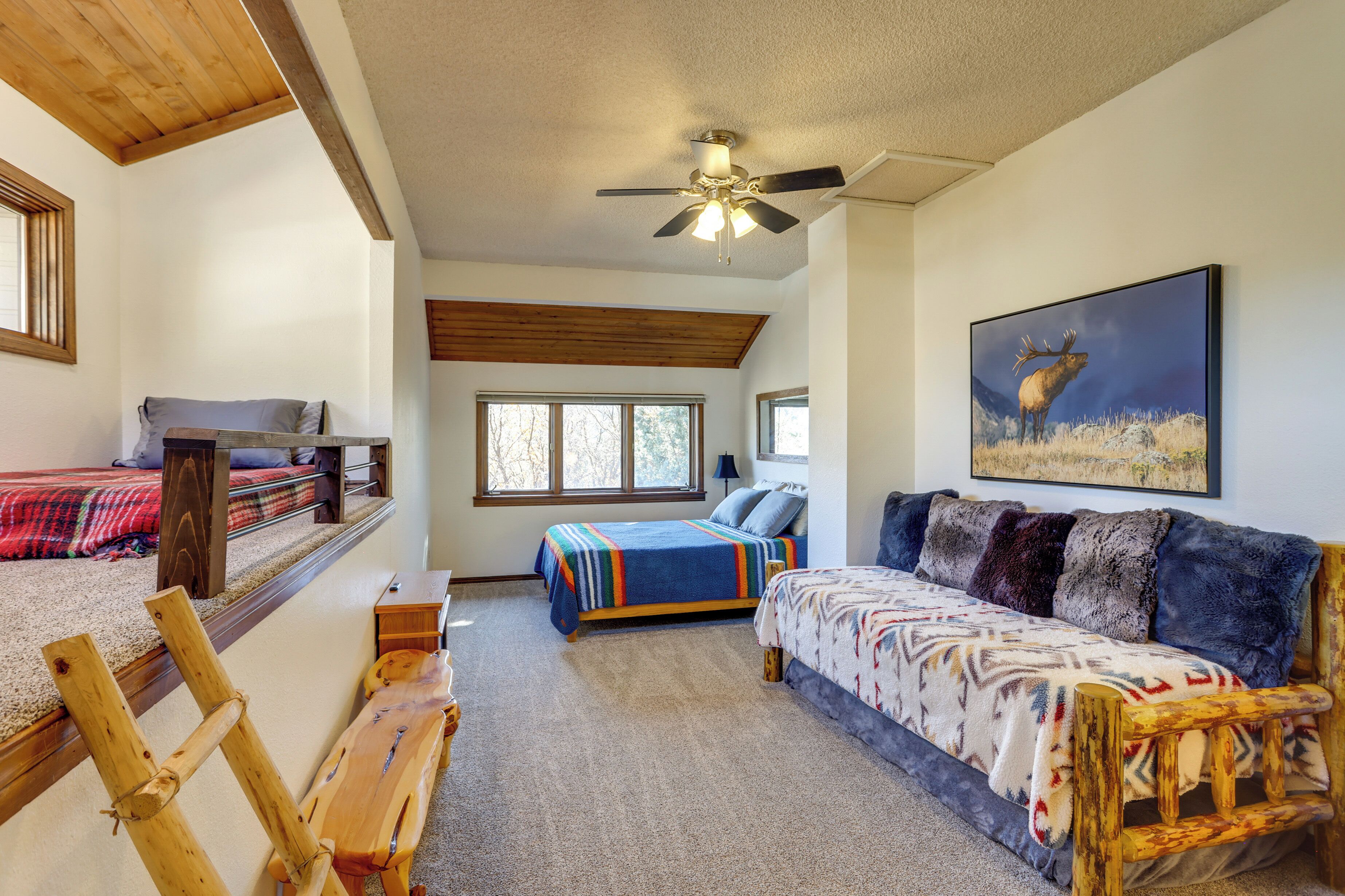 A bedroom with a bed, a sofa, a wooden bench, a ceiling fan, and a painting of an elk.