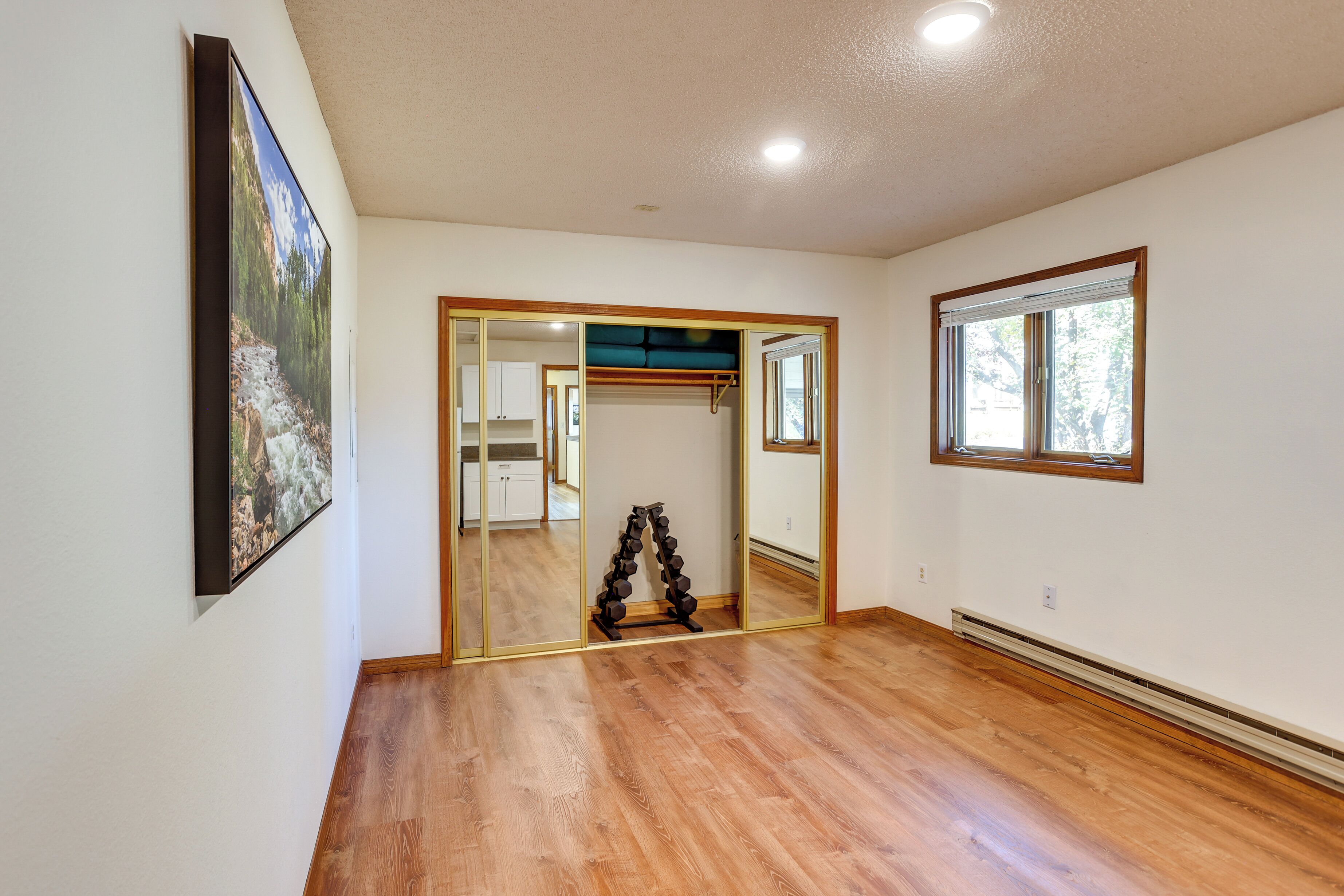 A spacious room with hardwood flooring, a large mirror, and a view of a kitchen through an open door.