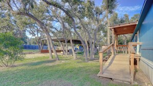 A wooden deck leading to a covered outdoor area with a blue building in the background. - Pet-friendly Canyon Lake Home ~ 3 Mi to Water! (Canyon Lake)