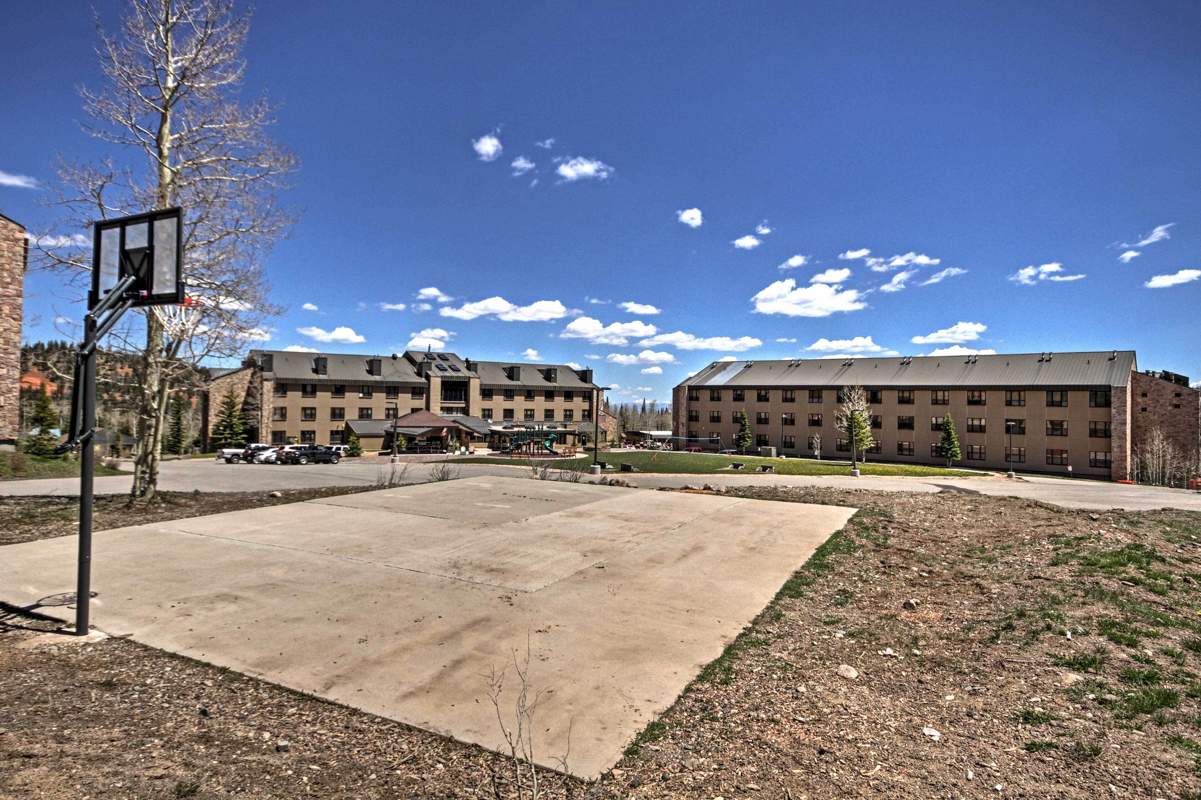 A basketball court with a hoop, surrounded by a building with multiple windows and a parking area.