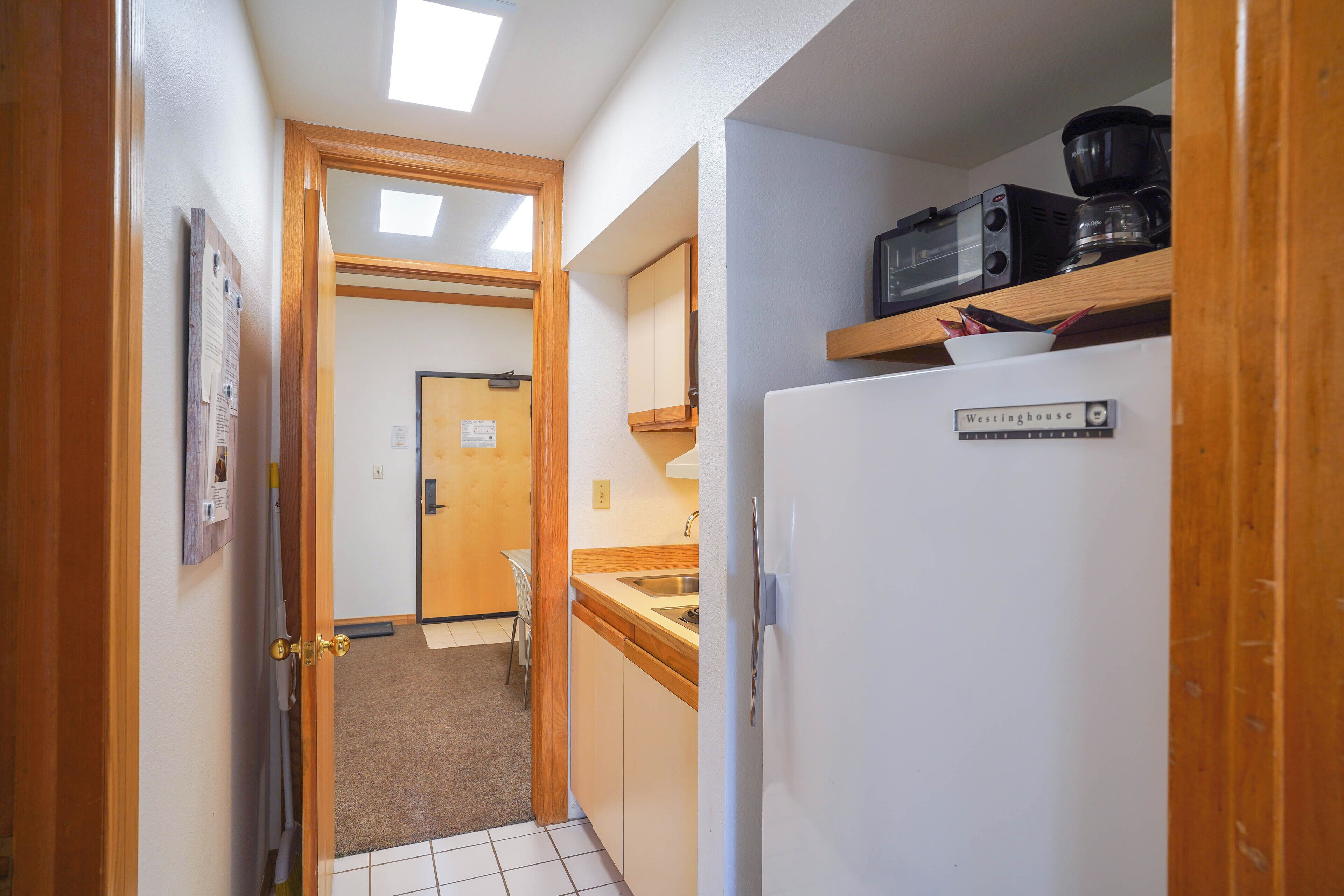 Un couloir étroit avec un réfrigérateur blanc, un cadre de porte en bois, un four à micro-ondes posé sur une étagère et un petit coin lavabo.