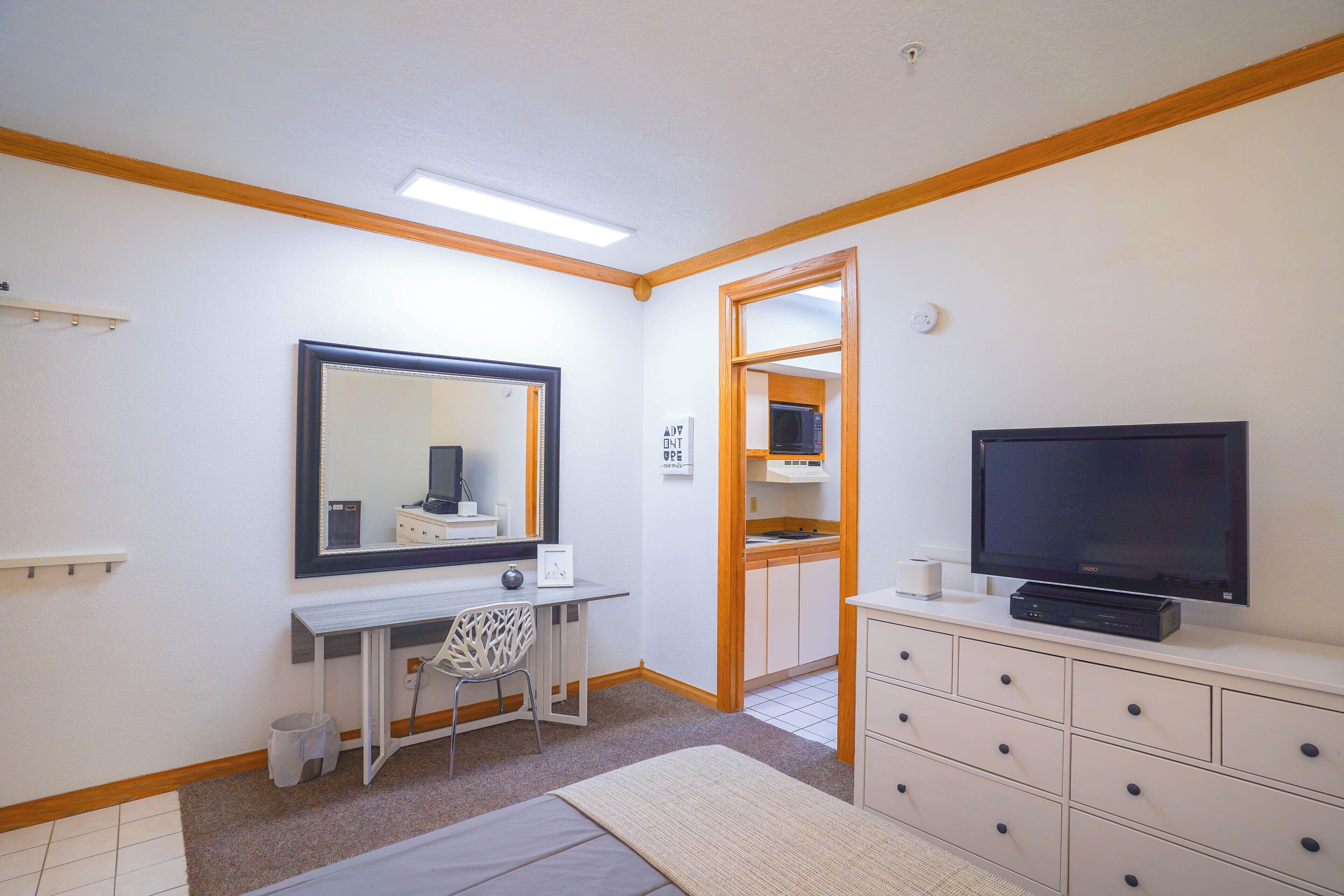 A compact hotel room with a kitchenette, a large mirror, a flat-screen TV, a desk with a chair, and a walk-in closet.