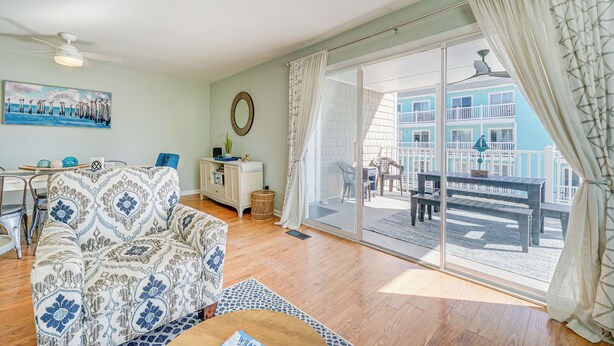 A living room with a patterned sofa, a coffee table, and a view of a balcony with outdoor furniture. - Steps to Ocean: Water-view Carolina Beach Unit! (Carolina Beach)